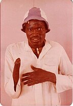 Shown is Garang Deng Bol, a Sudanese adult man wearing a white jalabiya layered over a T-shirt, with the T-shirt collar visible at the neckline. Bol is also wearing a striped hat with light and dark bands. Bol has a warm yet slightly hesitant expression. Bol's right arm is amputated just below the elbow, and their left hand is resting on their chest.