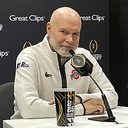 A football coach smiles in front of a microphone.