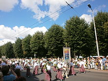 Seitliche Farbfotografie von einem Straßenumzug. Viele Frauen in rot-weißen Trachten und mit Sträußen in der Hand laufen mit einem Mann, der an der Spitze einen Stoffschild hält. An beiden Seiten stehen Zuschauer und im Hintergrund ist eine Baumallee.