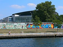 Frontale Farbfotografie von einem Flussufer mit einer Uferpromenade und einer langen Mauer mit Graffitis. Ein Mann sitzt links am Ufer und an der Mauer laufen einige Menschen. Rechts stehen vor und hinter der Mauer Bäume. Hinter der Mauer ist ein großes, modernes Glasgebäude.