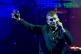 A middle-aged man standing in front of a coloured background with his right arm raised. He is holding a microphone in the opposite hand.