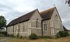 Three-quarter view of a flint and stone church in three sections. The middle section is the tallest and widest, and has a very large five-light lancet window with quatrefoils and an extremely narrow slit-like window above. The section on the left has a smaller lancet window and a similar slit. An extension on the right is mostly hidden.