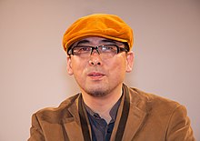 A bespectacled Tensai Okamura, in an orange corduroy cap