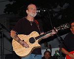 A man in his 50s or 60s, wearing a black top and jeans, with jewelry on both wrists and a crucifix around his neck. He is playing an acoustic guitar, and standing in front of a microphone. Other musicians are also visible.