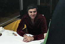 A Caucasian man wearing a dark burgundy shirt sits at a table.