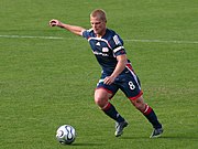 A soccer player in a New England Revolution kit is running and kicking the ball.