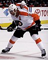 An ice hockey player is crouched over the ice, looking intently down at the floor. On his back a large '17' is visible with 'Carter' written above. His uniform is predominately white, with orange shoulders, and he is wearing a white helmet with a visor.