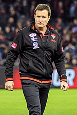 A man with light brown hair in a black jacket walks across a grassed playing field