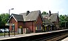 Two sets of rails between platforms beyond which is a brick building with two gables and a canopied area between them; a sign to the left says "Hough Green"