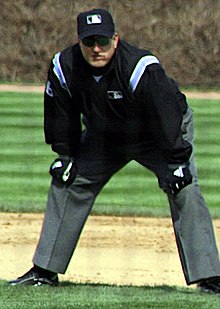 A baseball umpire crouching with his hands on his knees. He is wearing grey slacks, a black long sleeve jacket with baby blue stripes on the shoulders, black and white gloves, a black cap with the MLB logo on it, and sunglasses.