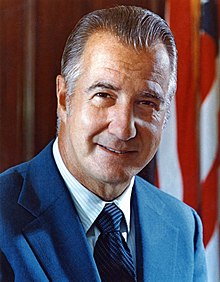 Spiro Agnew in 1972, a middle-aged white American male in suit and tie, standing in front of a furled flag
