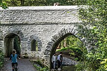 A bridge in St. Anne's Park. It is grey and with Arches of three sizes. There are trees in the background and a stream on the right side of the picture. A child is riding a bicycle, and three people are under one arch.