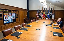 Photo of Biden seated alone at a table, looking at a videoconference screen