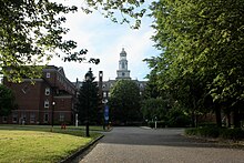 A red brick building featuring a white colonial tower. The building sits on a landscaped grounds populated with various trees, shrubs, and grass. A paved roadway navigates across the grounds and connects the building to Convent Road.