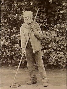 old bearded man wearing trousers and a coat, leaning on a pole