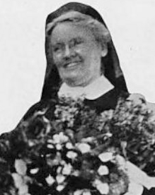 A smiling older white woman wearing a head covering, eyeglasses, and a high starched collar; there is a bouquet in the photo's foreground