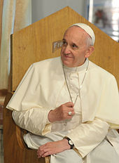 Frontale Farbfotografie vom Papst im weißen Papstgewand und Kappe, der lächelnd nach rechts blickt und auf einem Holzstuhl mit hoher Lehne sitzt. Er hält mit der rechten Hand seine silberne Halskette fest und trägt einen Siegelring und eine dunkle Armbanduhr.