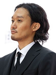 Fukunaga in three-quarter profile at press event wearing black suit and tie