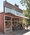 Photograph of a historic store, the Pioneer Emporium, in the Columbia Historic District. The building sports a tall false front, and several visitors stand in the shade of an awning.