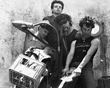 A band with five members. One on the left is shirtless and holding a saw, acting as if he was about to behead another member. A man stands in the back with an austere look. A fourth person is sticking out his tongue and the fifth is holding a guitar.