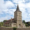 Three-quarter view of a flint church with a tall, three-stage tower topped with a dark grey spire. Low projections with paired arched windows flank the tower on both sides. A low flint wall curves round the front of the churchyard.