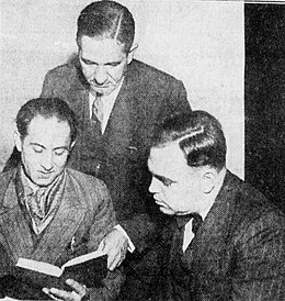 Black and white photo of Fard sitting next to two police officers. He is reading a book set in front of him.