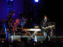 A rock band performing live onstage. On the left, a man is playing an bass guitar and a man is playing the drums. A man is singing and playing guitar in the center-right.