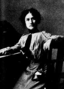 A young woman with fair skin and dark wavy hair, seated at a desk, wearing a striped shirtwaist and a skirt