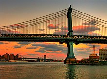 Zwei East-River-Brücken vom Wasser aus betrachtet. Vorne dominiert die Manhattan Bridge mit einem Hauptpylon das Bild, quer durchs Bild verläuft das doppelstöckige Brückendeck. Die charakteristischen, senkrecht von den geschwungenen Haupttragseilen zum Brückdeck verlaufenden Nebentragseile sind vor dem Hintergrund des Dämmerungshimmels deutlich zu erkennen. Im Bildhintergrund sind Uferbebauung und die Williamsburg Bridge mit ihren zwei Pylonen sichtbar.