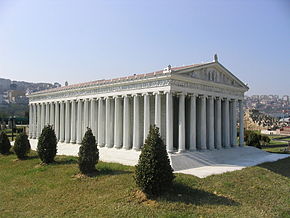 Artemis Tapınağı'nın maketi. Fotoğrafın tamamına hakim. Beyaz sütunlar üzerinde üçgen bir çatı, dikdörtgen bir yapı. Fotoğraf tapınağı ön sol çaprazdan görecek şekilde çekilmiş. Tapınağın ön cehpesi ve sol cephesi gözüküyor. Tapınak maketinin yanın minik ağaçlar dikilmiş, etrafta çimler var, hava açık.