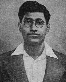 Young, clean-shaven face of a man with round spectacles and wavy black hair looking into the camera