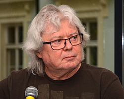 Vladimír Mišík wearing glasses, sitting behind microphone, looking left of camera