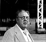 Albert R. Broccoli ist auf einer quadratischen schwarz-weiß Fotografie abgebildet. Er trägt eine Brille und hat wenig Haar. Er trägt einen hellen Anzug.