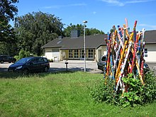 Auf der Wiese steht ein etwa drei Meter hohes, ungeordnetes Bündel armdicker Holzpfähle, die mit farbenfrohen Mustern bemalt sind. Das Arrangement erinnert an ein Bündel Mikadostäbe vor dem Fallenlassen.