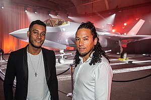 A photo of two men stood in front of a fighter jet on display.