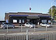 A red-bricked building with a blue sign reading "DAGENHAM EAST STATION" in white letters, three people walking in front, and three cars parked outside