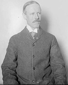 White man in medium shot. Man has receding hairline and mustache. He is wearing a collared shirt and a button down sweater or pinstriped jacket.