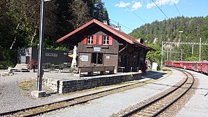 Two-story wooden building with gabled roof next to double-track railway line with red train on the far track