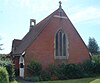 A red-brick church with a tiled roof extending nearly to ground level. Low shrubs surround the building on all sides. A three-light lancet window with stone mullions dominates the nearest side. A small stone cross and bell-tower are on the roof. The left-hand side has a white entrance porch.