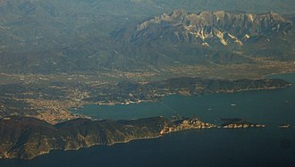 Unten im Bild das dunkelblaue Meer, dann im Vordergrund von links nach rechts eine Halbinsel mit Porto Venere rechts an der Spitze und einem Teil der Cinque-Terre-Küste links unten im Foto, dazwischen ein dunkelbrauner langgestreckter Hügelkamm mit Steilküste. In Bildmitte der Golf von La Spezia, die Stadt La Spezia links in der Bucht, rosabräunlich vermutlich wegen der Ziegeldächer. Oben ein Gebirgszug mit hellgrau wirkenden Bergspitzen vor allem im Bildteil rechts oben und einer Küstenlinie am Golf mit einem Vorgebirge, ebenfalls eine Halbinsel und einem dazwischen liegenden Tal, das Val di Magra ein wenig mehr zur Bildmitte hin