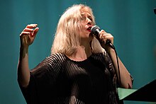 A blonde woman with a black formal dress singing on a microphone.