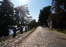 Frontale Farbfotografie von einer alten Straße mit Pflastersteinen. Links vom Ufer verdecken Bäume den Fluss und Steinbolzen führen entlang der Straße. Rechts steht zwischen Bäumen ein Haus.