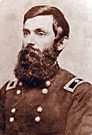 Black and white photo of a dark-haired man with a very bushy bread. He wears a dark military uniform with a brigadier general's star on his shoulder tabs.