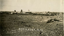 A black and white image of a rural town with three elevators and town buildings from an adjacent hay field