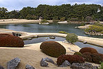 Garden with dried grassland, plants, woods and a large pond with islands.