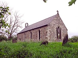 A photograph of a small stone church with open, glass-less windows. The church is surrounded by rough, uncultivated ground and the sky behind the church is bleak.