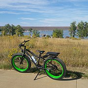 A black electric bicycle is resting on its kickstand on a sidewalk next to a grassy field leading to a small harbor. The bike has bright green wheel rims.
