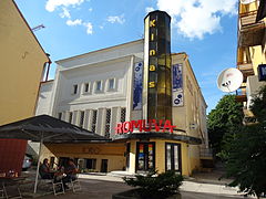 A white and yellow building with an black and yellow multi-sided tower on the roof at the front. It says "Kinas" in yellow. Below it is the word "Romuva" in red.