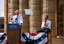 Photo of Biden outdoors behind a lectern, with Obama seated behind him and smiling