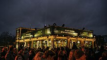 Small building with glass walls, with signs on top and spectators outside